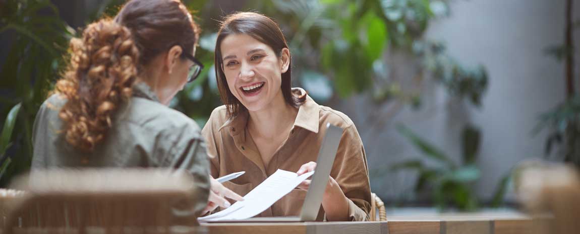 Två kvinnor samtalar vid ett bord med en laptop